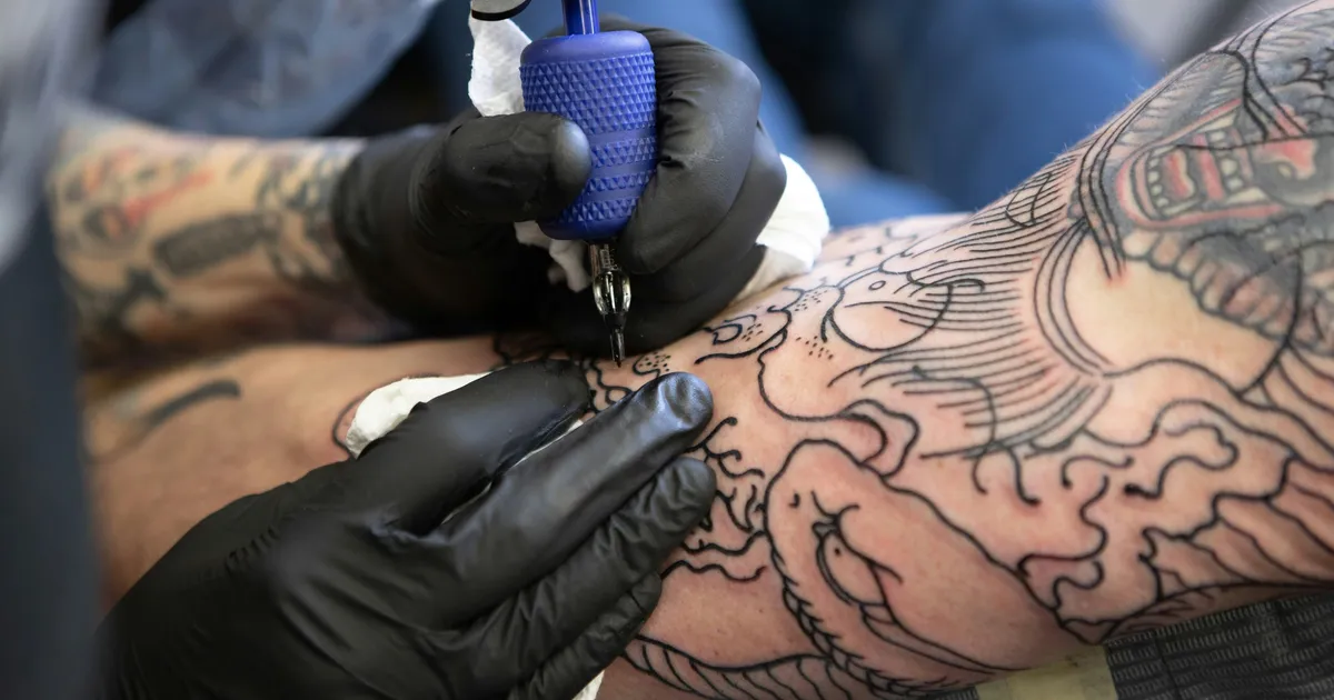 Close-up of a tattoo artist's gloved hands using a tattoo machine to add linework to a heavily tattooed forearm